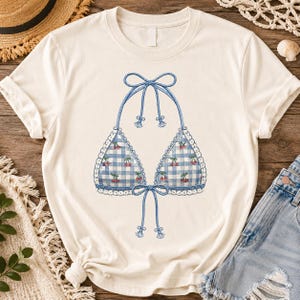 Puede incluir: Una camiseta de manga corta color crema con un gráfico central de un top de bikini de cuadros vichy azul y blanco con estampado de cerezas. El diseño incluye lazos azules tipo cuerda en el cuello y el centro, con un borde de encaje blanco.