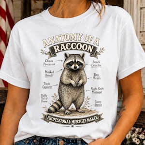 May include: A white t-shirt featuring a graphic illustration of a raccoon in the center. The text reads ANATOMY OF A RACCOON, PROFESSIONAL MISCHIEF MAKER, Chaos Processor, Masked Bandit, Trash Explorer, Fluffy Tail of Doom, Snack Detector, Tiny Hands, Night Shift Manager, and Sassy Stance.