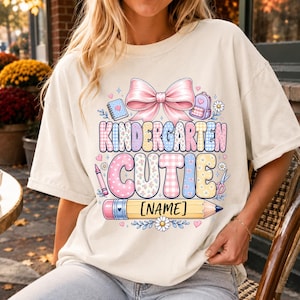 May include: A cream-colored t-shirt featuring a graphic print with a large pink bow, a notebook, a backpack, and the text "KINDERGARTEN CUTIE" in colorful patterned letters. Below the text is a yellow pencil graphic with the placeholder "[NAME]" inside.