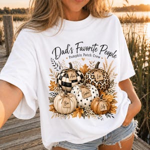 Peut inclure: Un t-shirt blanc avec un motif de cinq citrouilles aux motifs variés, notamment à carreaux, léopard et à pois. Chaque citrouille porte une étiquette avec les prénoms Ava, Leo, Ben, Sophie et Lily. Le texte indique Dad's Favorite People Pumpkin Patch Crew.