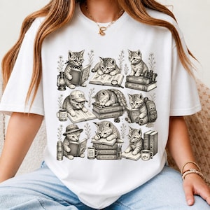 May include: White t-shirt featuring a detailed illustration of several kittens reading books. The kittens are depicted in various poses, some wearing hats or glasses, with books titled "USA", "Natural History", and "Field Notes".