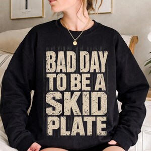 Puede incluir: Sudadera negra con el texto "BAD DAY TO BE A SKID PLATE" en una fuente desgastada de color blanquecino. El diseño está centrado en el pecho. La sudadera es de color negro liso.