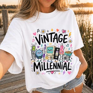 以下が含まれることがあります： 黒で「VINTAGE MILLENNIAL」と書かれたカラフルなグラフィックプリントが施された白いTシャツ。デザインには、折りたたみ式携帯電話、CDプレーヤー、ペン、コンピューター、VHSテープ、ラバランプ、シリアルボウル、カメラの漫画風イラストが含まれています。