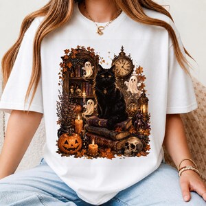 May include: A white t-shirt featuring a graphic print of a black cat sitting on a stack of old books. The scene includes a carved pumpkin, lit candles, small ghosts, a skull, a wall clock, and autumn leaves, creating a detailed Halloween-themed illustration.