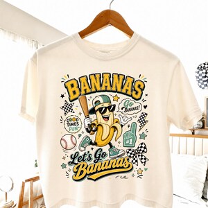 May include: Cream-colored t-shirt featuring a cartoon banana wearing sunglasses, a baseball cap, and sneakers, holding a baseball bat. The shirt has the text "BANANAS" and "Let's Go Bananas" with baseball-themed graphics.