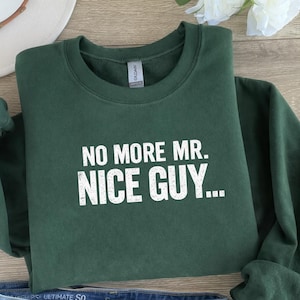 May include: A dark green sweatshirt with the text "NO MORE MR. NICE GUY..." in white block letters. The sweatshirt is folded and appears to be made of a soft material. The Gildan brand tag is visible.