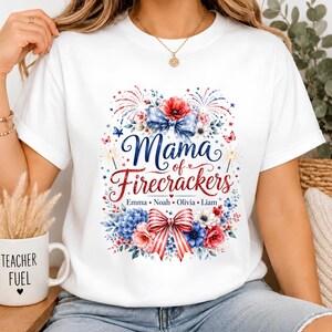 Puede incluir: Una camiseta blanca de manga corta con un estampado gráfico que incluye el texto "Mama of Firecrackers" en letra cursiva azul y roja. El diseño presenta flores rojas y azules, fuegos artificiales y un lazo a rayas, con los nombres Emma, Noah, Olivia y Liam.