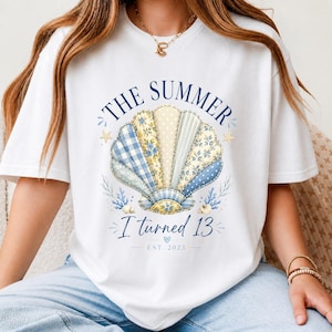 Könnte beinhalten: Ein weißes T-Shirt mit einer großen Grafik in der Mitte, die eine Muschel aus verschiedenen blau-gelb gemusterten Stoffsegmenten zeigt. Der Text lautet THE SUMMER I turned 13 EST. 2023, ergänzt durch kleine Seesterne, Korallen und Muscheln.