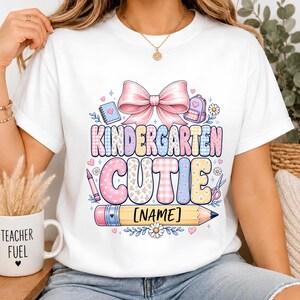 May include: A white t-shirt featuring a colorful graphic with the text KINDERGARTEN CUTIE and [NAME] on a yellow pencil. The design includes a large pink bow, a notebook, a backpack, a crayon, scissors, and small flowers in pastel tones.