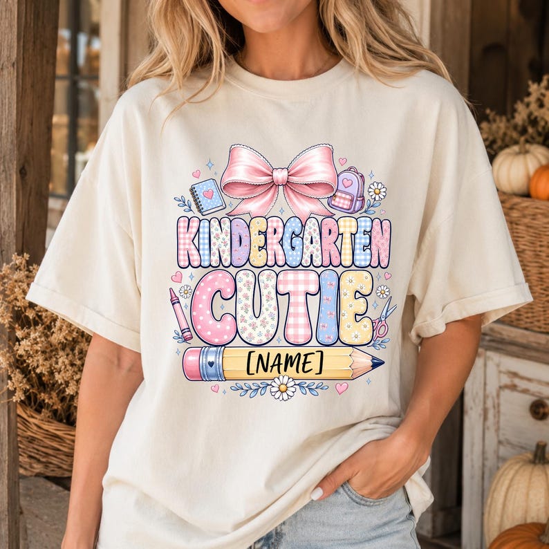 May include: A cream-colored t-shirt featuring a graphic with the text KINDERGARTEN CUTIE and [NAME] on a yellow pencil. The design includes a large pink bow, a notebook, a backpack, a crayon, scissors, and small flowers in pastel colors.