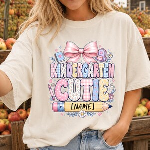 May include: A cream-colored t-shirt featuring a graphic with a large pink bow, a notebook, a backpack, and a pencil. The text reads KINDERGARTEN CUTIE and [NAME] on the pencil. The design includes pastel-colored patterns, flowers, and hearts.