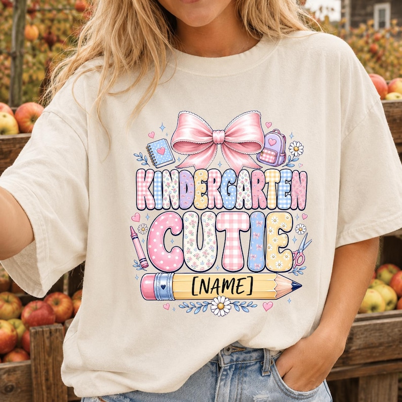 May include: A cream-colored t-shirt featuring a graphic with a large pink bow, a notebook, a backpack, and a pencil. The text reads KINDERGARTEN CUTIE and [NAME] on the pencil. The design includes pastel-colored patterns, flowers, and hearts.