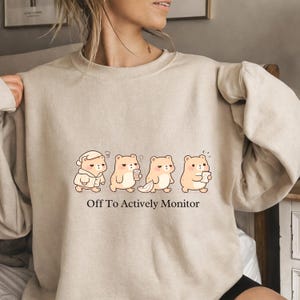 Puede incluir: Sudadera beige con una fila de osos de dibujos animados en varios estados de vigilia. Los osos son de color marrón claro con ojos negros y se representan con una máscara para dormir, café y un bloc de notas. El texto "Off To Actively Monitor" está impreso debajo.