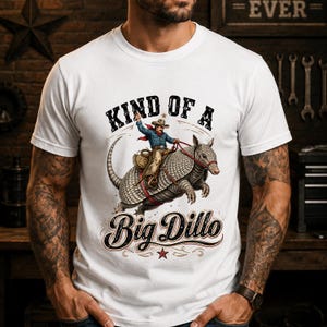 May include: A white short-sleeved t-shirt featuring a graphic of a cowboy riding an armadillo. The text above reads "KIND OF A" and the text below reads "Big Dillo" in a stylized font. The design includes a small red star at the bottom.