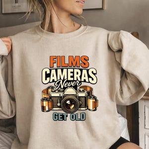 Op de afbeelding: Beige sweatshirt met een grafisch ontwerp met een vintage camera, filmrollen en de tekst "FILMS CAMERAS Never GET OLD" in oranje, zwart en blauw. Het ontwerp heeft een retro esthetiek, perfect voor fotografie liefhebbers.