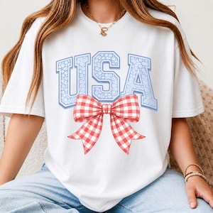 Puede incluir: Camiseta blanca con la palabra "USA" en letras azul claro con estampado floral. Un gran lazo de cuadros rojos y blancos está centrado debajo del texto. La camiseta es un diseño casual y patriótico.