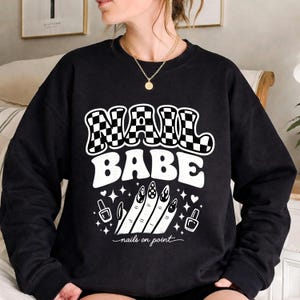 Könnte beinhalten: Schwarzes Sweatshirt mit dem Schriftzug "NAIL BABE" in einem Retro-Stil, wobei das Wort "NAIL" ein Schachbrettmuster aufweist. Das Design umfasst Illustrationen von Nagellackflaschen, Maniküre-Nägeln und den Satz "nails on point".