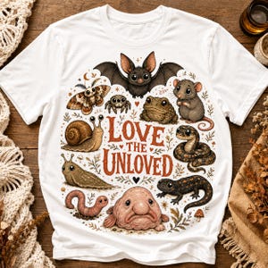 May include: A white short-sleeve t-shirt featuring a colorful graphic of various animals including a bat, moth, spider, toad, mouse, snake, snail, slug, worm, and blobfish. The text "LOVE THE UNLOVED" is printed in the center in a rustic red font.