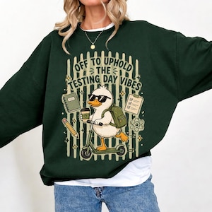Può includere: Felpa verde scuro con grafica di un'anatra con occhiali da sole, su uno scooter, con uno zaino e un caffè. Il testo recita: "OFF TO UPHOLD THE TESTING DAY VIBES".