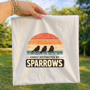 May include: Off-white square fabric with a retro design featuring three black birds perched on a line, set against a sunset of orange, yellow, and blue stripes. The text reads "EASILY DISTRACTED BY SPARROWS."