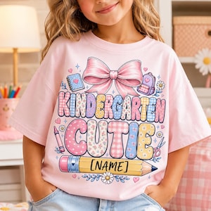 May include: A light pink t-shirt featuring a large graphic with a pink bow, a notebook, a backpack, a crayon, and scissors. The text reads KINDERGARTEN CUTIE with a yellow pencil graphic at the bottom containing the placeholder text [NAME].