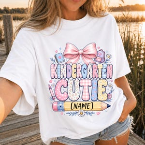 May include: A white t-shirt featuring a graphic print with a large pink bow above the text KINDERGARTEN CUTIE in colorful patterned letters. Below the text is a yellow pencil with the placeholder text NAME, surrounded by small school icons.