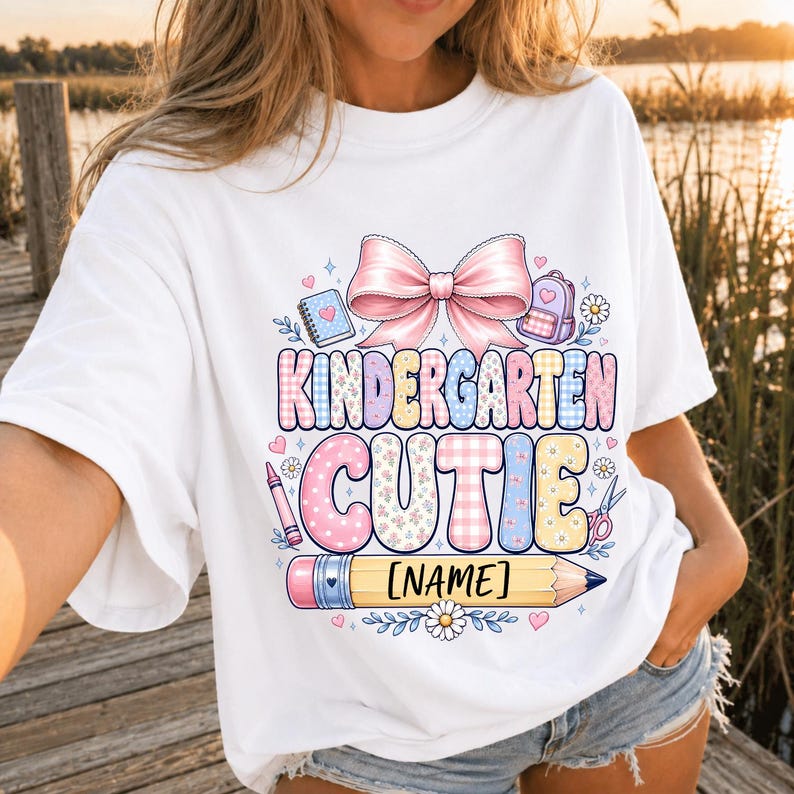 May include: A white t-shirt featuring a graphic print with a large pink bow above the text KINDERGARTEN CUTIE in colorful patterned letters. Below the text is a yellow pencil with the placeholder text NAME, surrounded by small school icons.