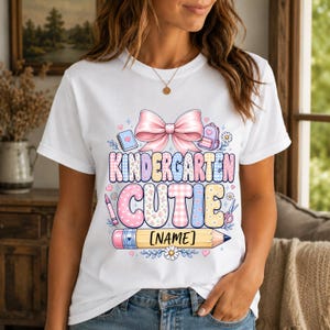 May include: A white t-shirt featuring a graphic print with the text KINDERGARTEN CUTIE and [NAME] on a yellow pencil. The design includes a large pink bow, a notebook, a backpack, a crayon, scissors, and small flowers in pastel colors.