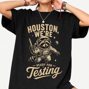 Könnte beinhalten: Schwarzes T-Shirt mit einem Waschbär-Astronauten, der einen Bleistift hält. Der Text "Houston, We're Ready for Testing" ist im Vintage-Stil gehalten. Der Waschbär trägt einen Raumanzug, mit Sternen und einem Planeten im Hintergrund.