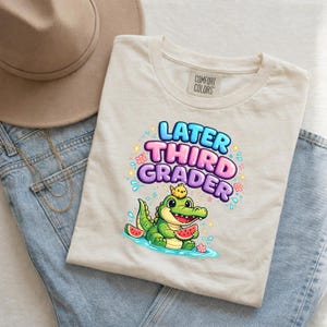 Könnte beinhalten: Beiges Comfort Colors T-Shirt mit farbenfroher Grafik. Die Grafik zeigt "Later Third Grader" über einer Illustration eines grünen Alligators, der Wassermelone hält. Das T-Shirt liegt auf einer blauen Jeans und einem beigen Hut.