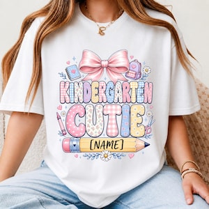 May include: A white t-shirt featuring a graphic with a large pink bow above the text KINDERGARTEN CUTIE in pastel plaid and floral patterns. The design includes a pencil with the text [NAME], a notebook, a backpack, a crayon, and scissors.