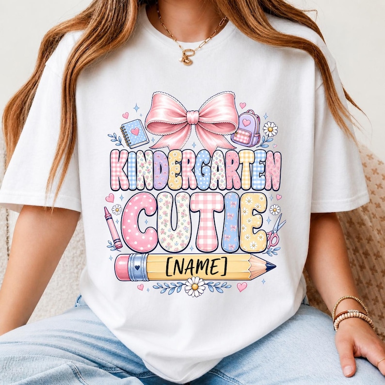 May include: A white t-shirt featuring a graphic with a large pink bow above the text KINDERGARTEN CUTIE in pastel plaid and floral patterns. The design includes a pencil with the text [NAME], a notebook, a backpack, a crayon, and scissors.