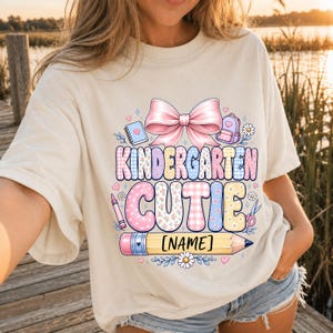 May include: A cream-colored t-shirt featuring a graphic print with a large pink bow, school supplies, and the text "KINDERGARTEN CUTIE" above a yellow pencil graphic with a placeholder for a name. The design includes floral and plaid patterns.