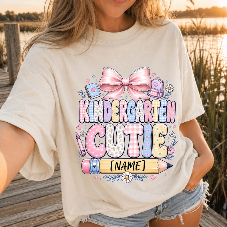 May include: A cream-colored t-shirt featuring a graphic print with a large pink bow, school supplies, and the text "KINDERGARTEN CUTIE" above a yellow pencil graphic with a placeholder for a name. The design includes floral and plaid patterns.