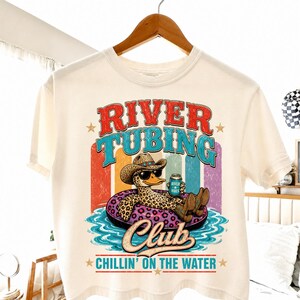 Peut inclure: T-shirt crème avec un canard de dessin animé portant un chapeau de cow-boy et des lunettes de soleil, flottant dans une bouée léopard. Le t-shirt affiche les mots "River Tubing Club" et "Chillin' on the Water".