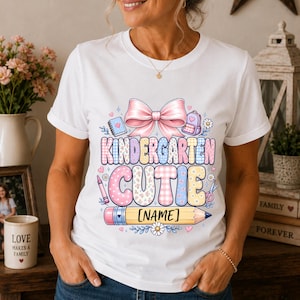 May include: A white short-sleeved t-shirt featuring a graphic print with a large pink bow, school supplies, and the text "KINDERGARTEN CUTIE" above a yellow pencil graphic with the placeholder text "[NAME]" in black brackets.