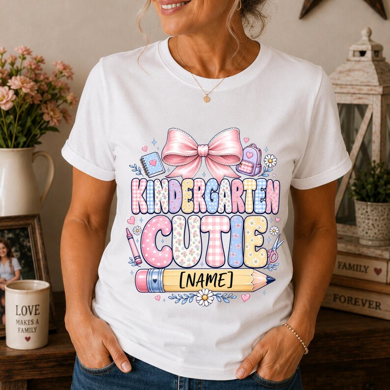 May include: A white short-sleeved t-shirt featuring a graphic print with a large pink bow, school supplies, and the text "KINDERGARTEN CUTIE" above a yellow pencil graphic with the placeholder text "[NAME]" in black brackets.