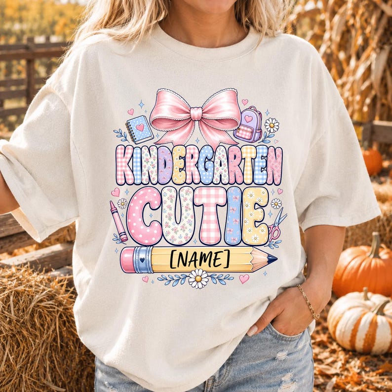 May include: A cream-colored t-shirt featuring a graphic print with a large pink bow, a notebook, a backpack, and a pencil. The text reads KINDERGARTEN CUTIE and [NAME]. The design includes pastel-colored patterns, small hearts, and flowers.