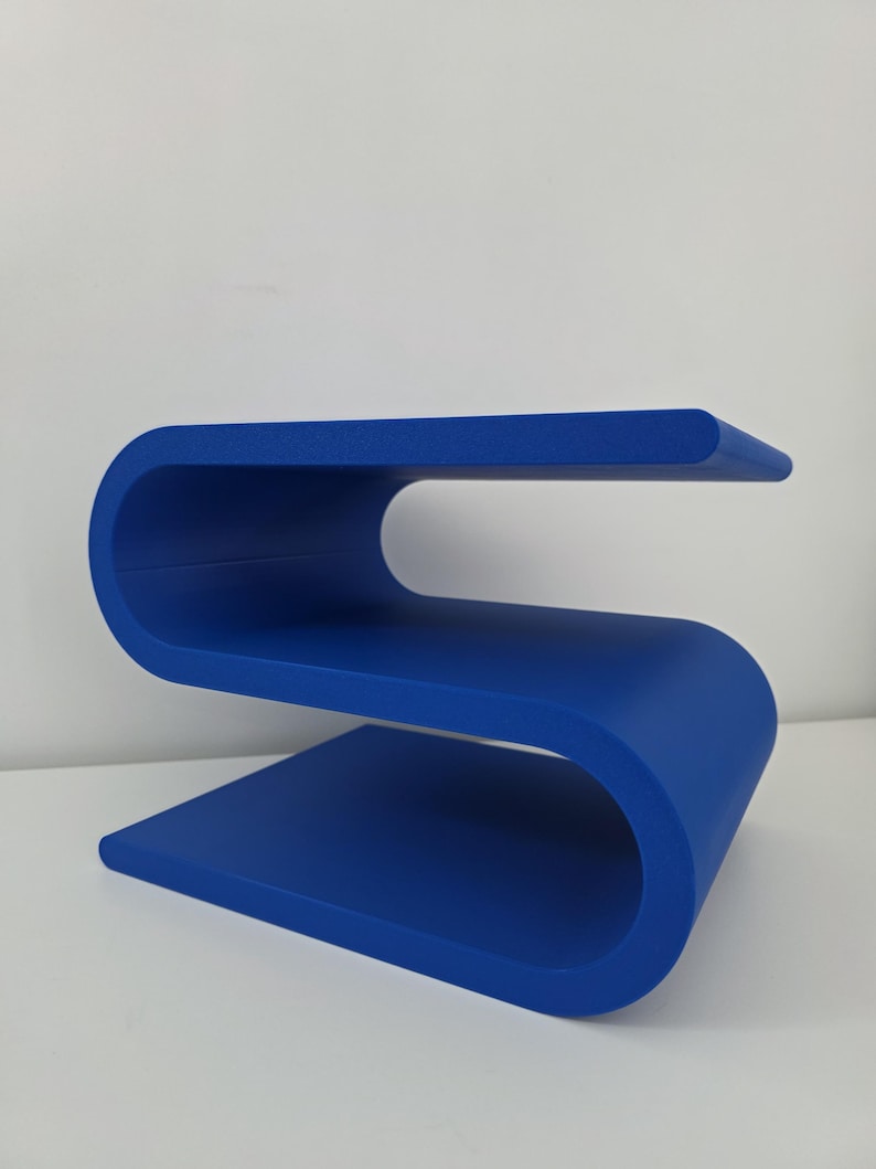 May include: A vibrant blue side table featuring a continuous S-shaped design with two horizontal surfaces. The structure is formed from a single piece of material that curves to create an upper shelf and a lower base, providing a sleek profile.
