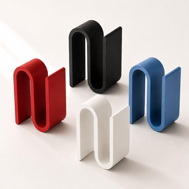 May include: Four S-shaped holders in red, black, white, and blue. Each holder features a continuous, curved design with a flat base and two vertical sides, creating a channel for storage. The objects are arranged in a cluster on a plain surface.