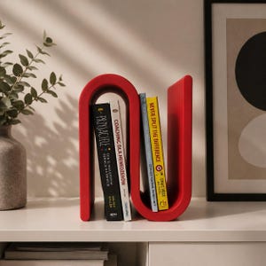 May include: A bright red, S-shaped magazine rack or book holder with a smooth, curved design. It holds four books upright, including titles like Przyjaciele, Coaching dla Menedżer&oacute;w, and Never Split the Difference by Chris Voss, organized in two sections.