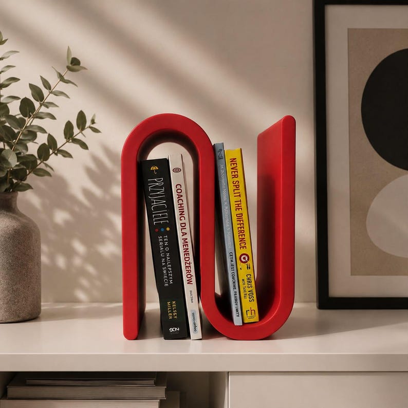 May include: A bright red, S-shaped magazine rack or book holder with a smooth, curved design. It holds four books upright, including titles like Przyjaciele, Coaching dla Menedżer&oacute;w, and Never Split the Difference by Chris Voss, organized in two sections.