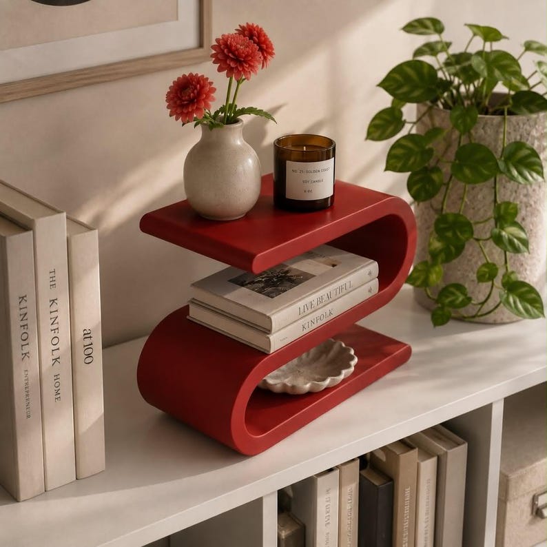 May include: A red S-shaped side table holding a cream-colored vase with two red flowers, a glass candle, and a book titled Live Beautiful. The table is positioned on a white surface next to three books and a potted green plant in a textured pot.