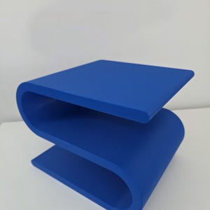 May include: A solid blue side table featuring a continuous S-shaped design. The piece is constructed from a single piece of material with smooth, rounded edges and a flat top surface, creating a distinct serpentine profile that rests on the floor.