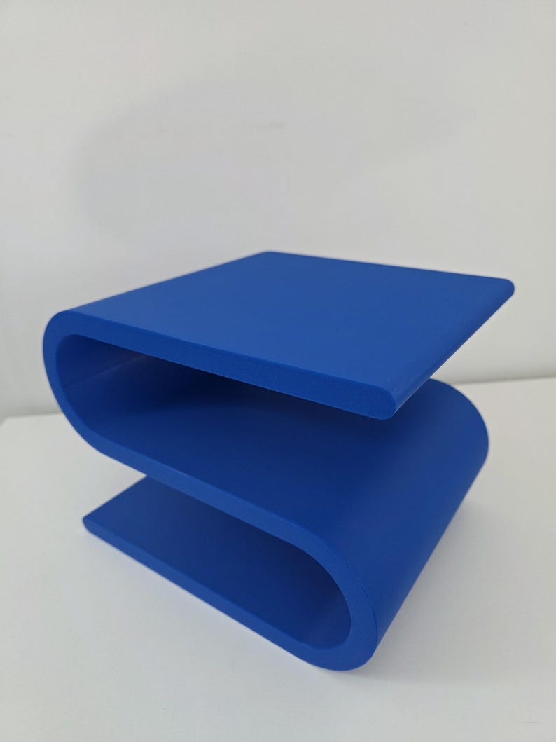 May include: A solid blue side table featuring a continuous S-shaped design. The piece is constructed from a single piece of material with smooth, rounded edges and a flat top surface, creating a distinct serpentine profile that rests on the floor.