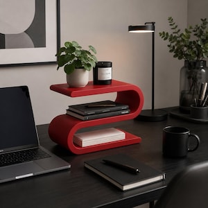 May include: A bright red, S-shaped desk organizer with three horizontal tiers. The top tier holds a small potted green plant and a black candle. The middle tier contains two stacked black books, and the bottom tier holds a stack of white paper.