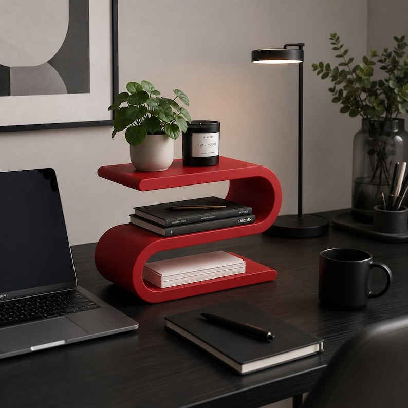 May include: A bright red, S-shaped desk organizer with three horizontal tiers. The top tier holds a small potted green plant and a black candle. The middle tier contains two stacked black books, and the bottom tier holds a stack of white paper.