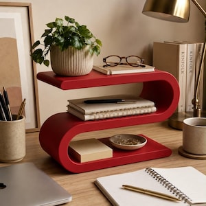 May include: A red S-shaped desk organizer with three tiers. The top tier holds a potted green plant and brown-rimmed glasses on a notebook. The middle tier holds a spiral-bound notebook and a pen. The bottom tier holds a small notepad and a bowl.