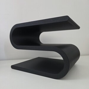 May include: A matte black side table featuring a continuous S-shaped design with a flat base, a curved middle section, and a flat top surface. The structure is formed from a single piece of material, creating a sleek, fluid, and geometric silhouette.