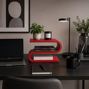 May include: A bright red S-shaped wooden shelf with three horizontal tiers. The top tier holds a small green potted plant and a black candle. The middle tier contains two stacked black books. The bottom tier holds a stack of white paper.